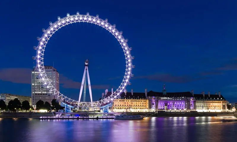An observation wheel lit white and violet above a river at night, each of its capsules a separate point of light